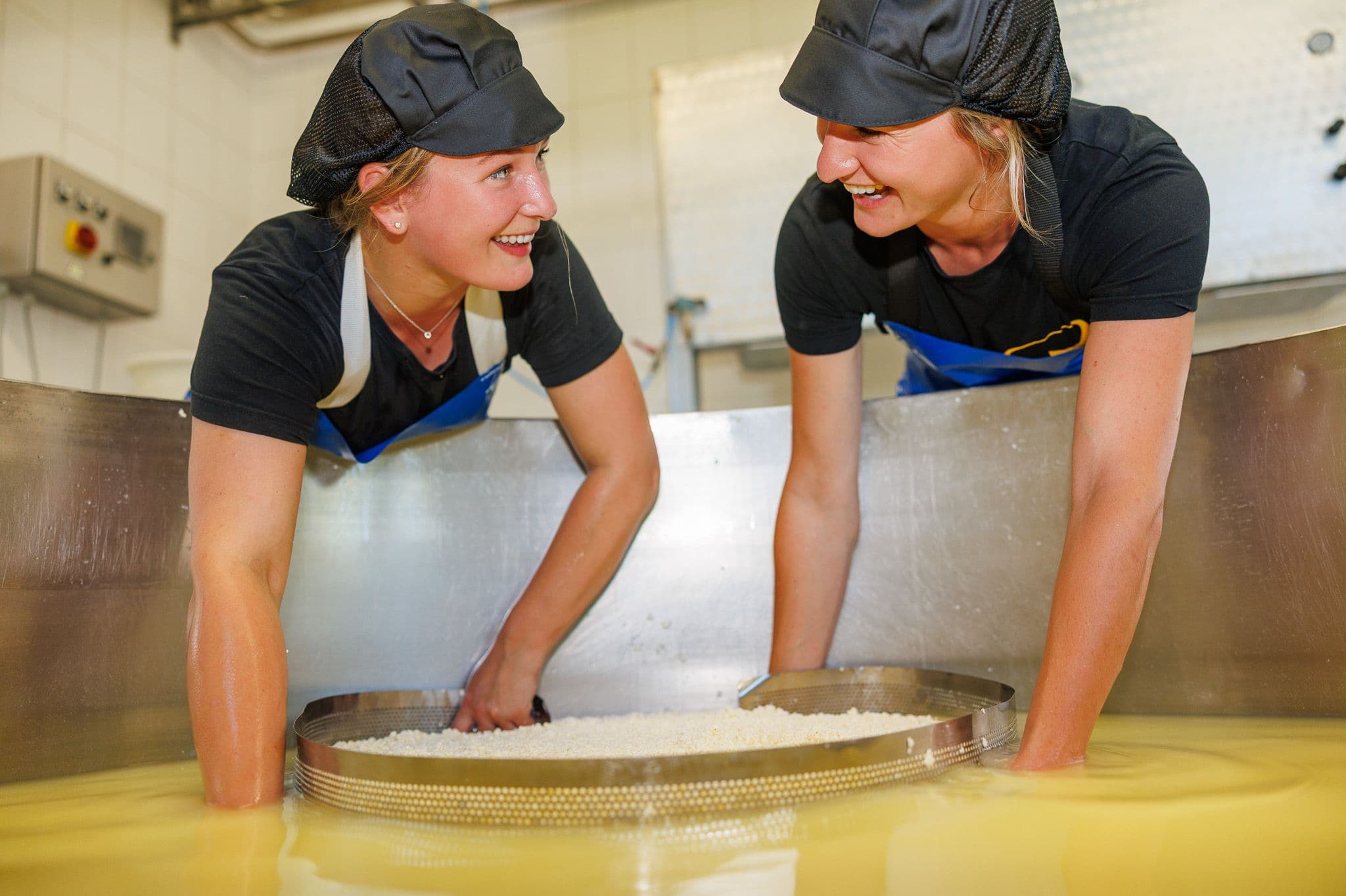 Twee jonge kaasmakers scheppen wrongel met een zweef uit de kaastobbe.